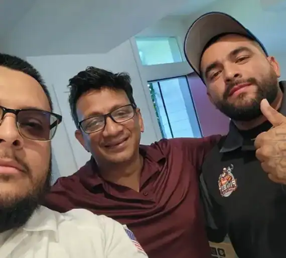Three men pose for a selfie indoors; one is giving a thumbs up, and all appear to be smiling slightly.